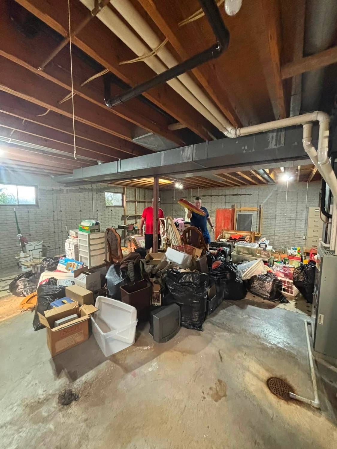 Unfinished basement crowded with bags, boxes, furniture and an old television while two crew members sort items