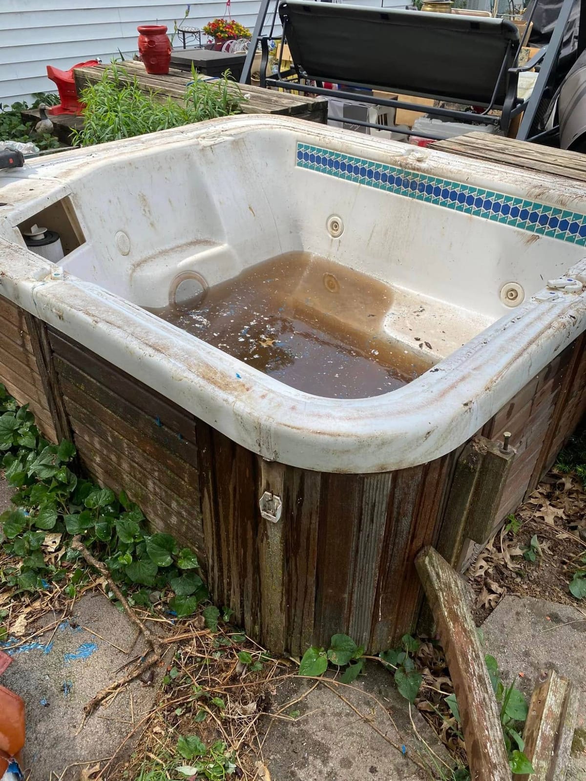 Old hot tub with a weathered wood surround, half full of murky water, beside a backyard deck